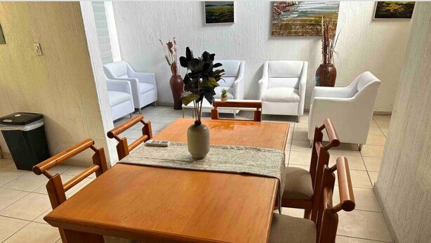 A dining area with a wooden table and chairs, a vase with plants, and white sofas in the background.