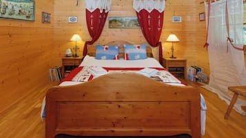 A wooden cabin bedroom with a large bed, two bedside tables, a nightstand, a window with curtains, and a painting on the wall.