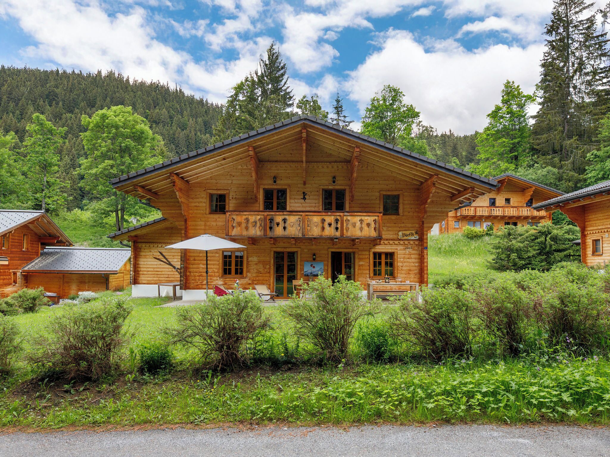 A wooden chalet with a balcony, surrounded by lush greenery and a mountainous backdrop.