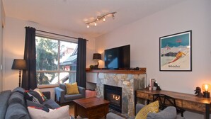 A cozy living room with a fireplace, comfortable seating, a flat-screen TV, and a scenic poster. - Quiet tranquil setting. Updated townhouse Free parking, walk to Village and shops, common hot tub, (Whistler)