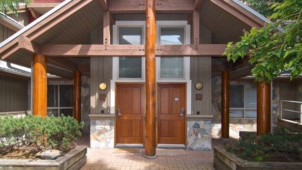 A two-story building with a symmetrical facade, featuring a central entrance with double doors, flanked by stone pillars and wooden beams. - Quiet tranquil setting. Updated townhouse Free parking, walk to Village and shops, common hot tub, (Whistler)