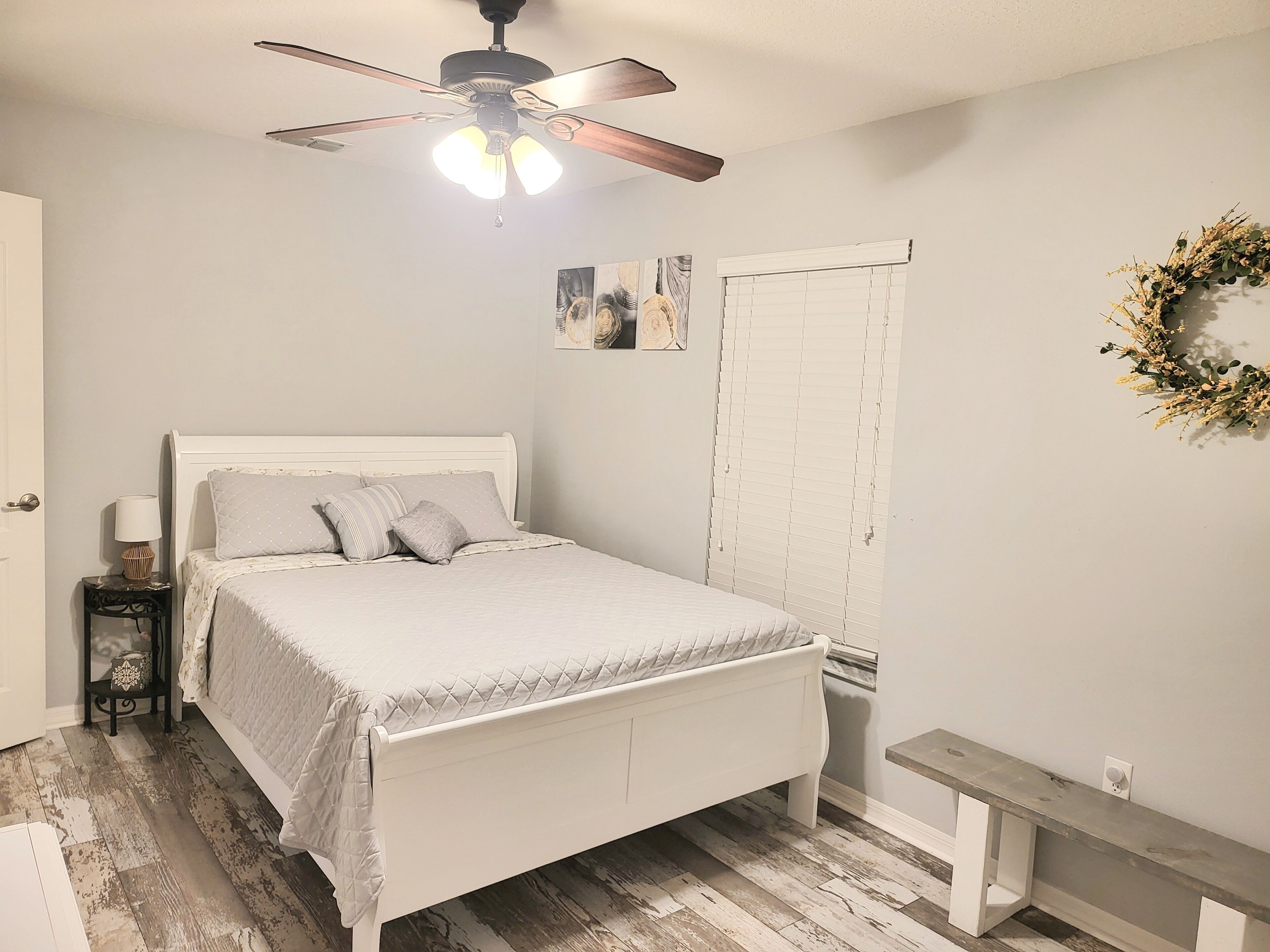 A bedroom with a bed, a ceiling fan, a bedside table with a lamp, a bench, and a wall-mounted wreath.