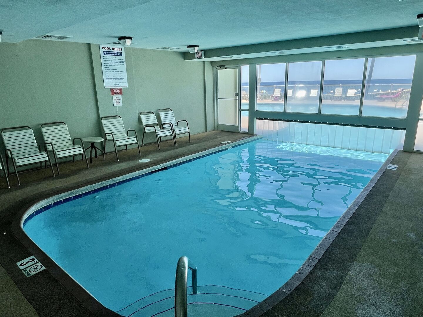 Piscina interna com paredes de vidro transparente, área de estar junto à piscina e vista para a praia.