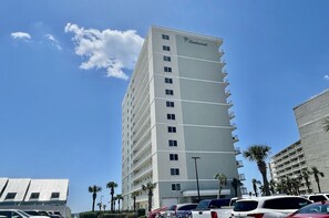 Ein hohes, mehrstöckiges Hotel mit Balkonen auf jeder Etage, gelegen in einer sonnigen Gegend mit Palmen und parkenden Autos.