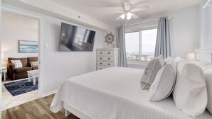 A bedroom with a large bed, a ceiling fan, a mounted TV, a dresser, and a window with curtains.