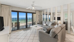 Un vaste espace de vie avec une grande porte-fenêtre coulissante donnant sur un balcon avec vue sur l’océan. On y trouve un canapé gris, une table basse blanche et un téléviseur à écran plat posé sur un meuble.