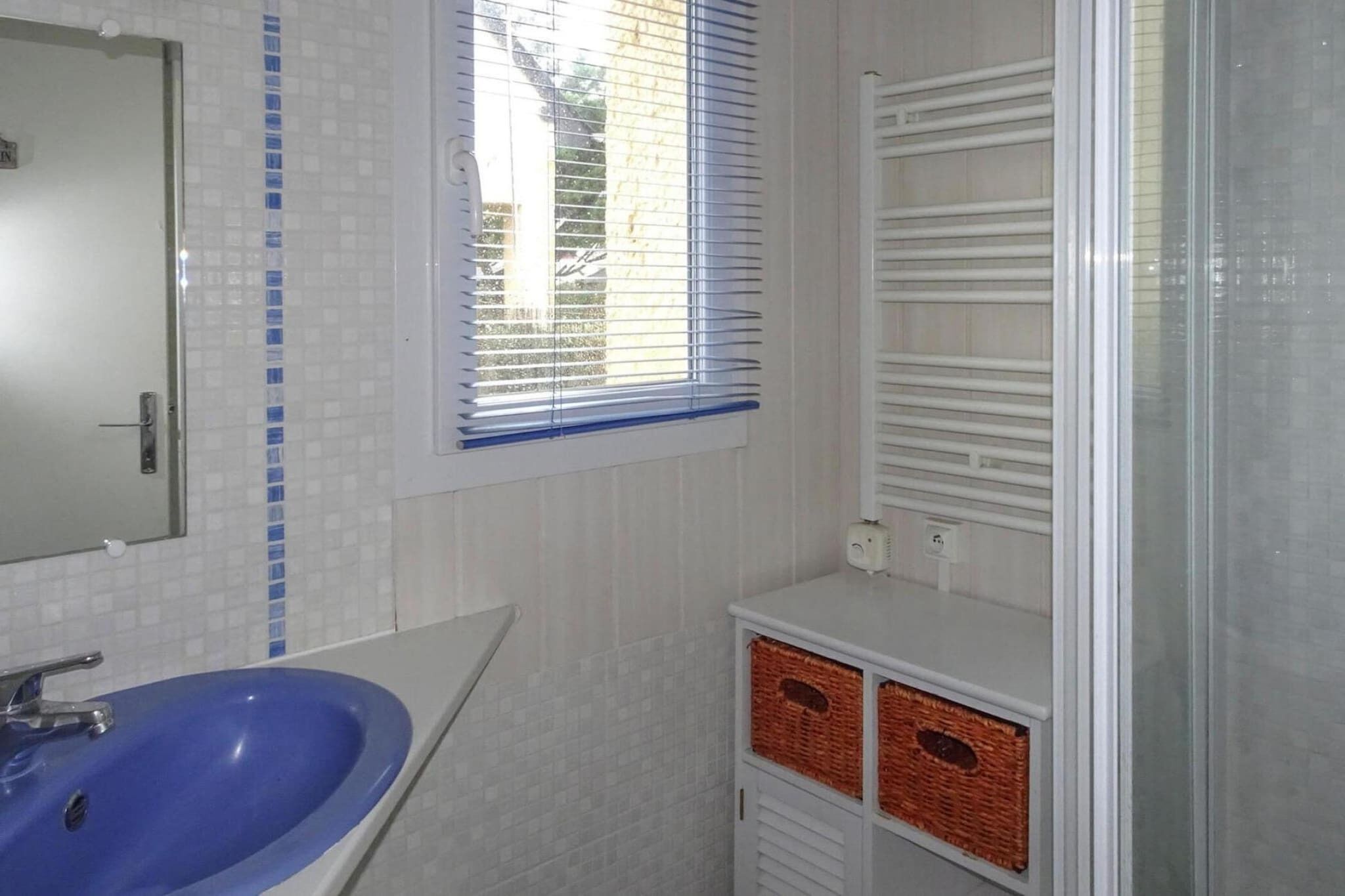 A bathroom with a blue sink, a mirror, a window with blinds, and a white cabinet with baskets.