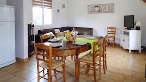 Zona pranzo con tavolo in legno apparecchiato per quattro persone, circondato da sedie in legno. Sullo sfondo, si vedono una televisione e un mobile.