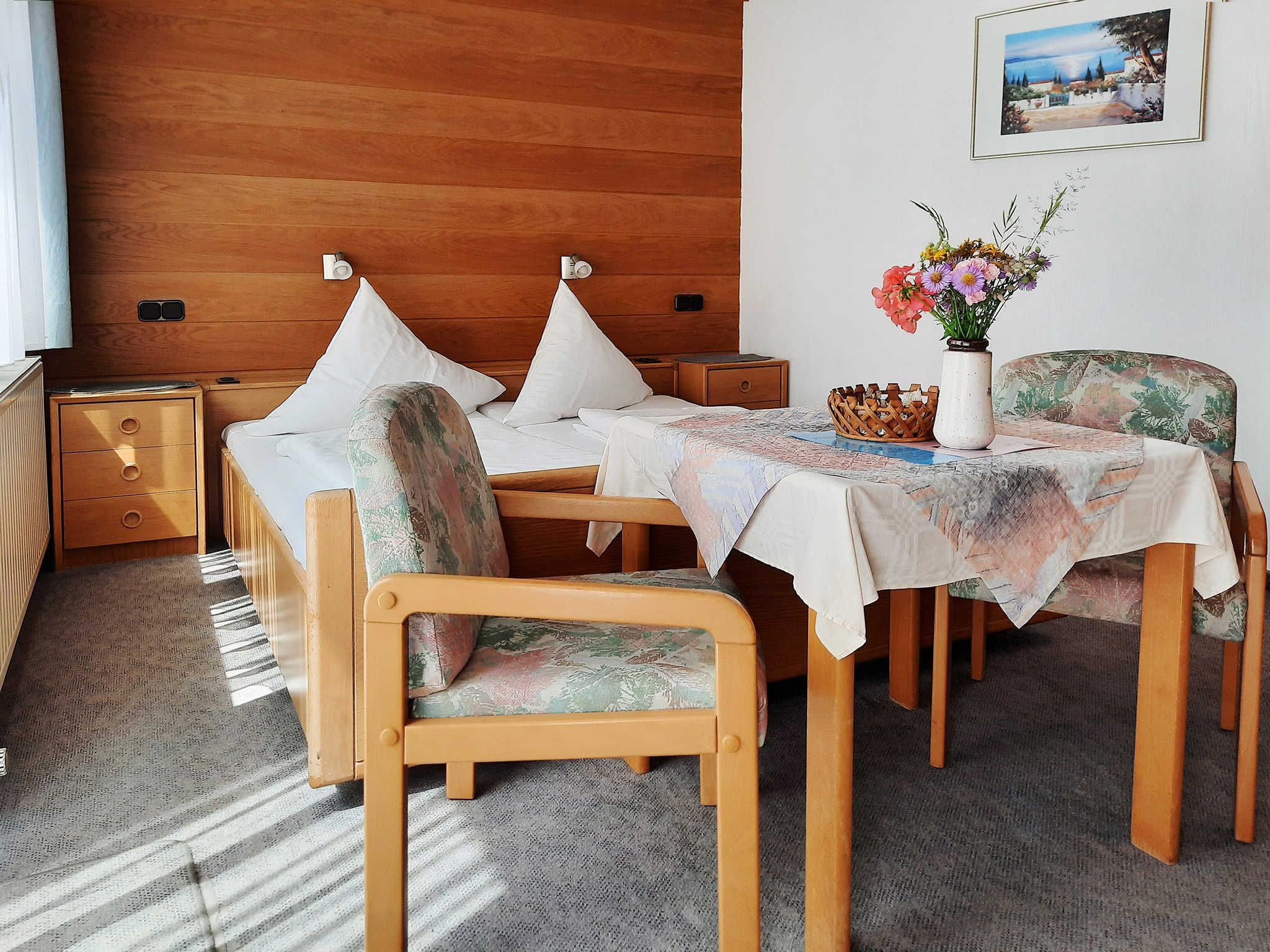 A hotel room with a wooden headboard, a bed with white linens, a small table with a floral tablecloth, a chair with a floral pattern, and a wall-mounted picture.