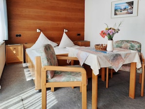 Habitación de hotel con cabecera de madera, cama con ropa blanca, mesita con mantel floral, silla con estampado floral y un cuadro colgado en la pared.