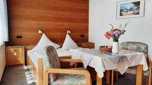 A hotel room with a wooden headboard, a bed with white linens, a small table with a floral tablecloth, a chair with a floral pattern, and a wall-mounted picture. - Café-Pension Krems - Double room (Waischenfeld-Heroldsberg-Tal)