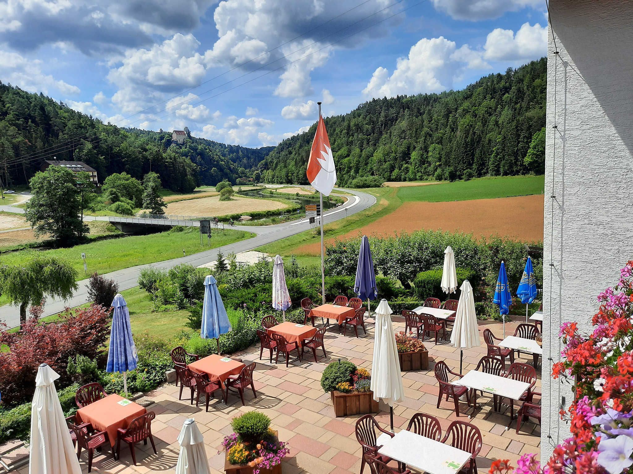 Eine Terrasse mit Sitzgelegenheiten im Freien, einer Flagge und Blick auf eine Straße und einen Wald.