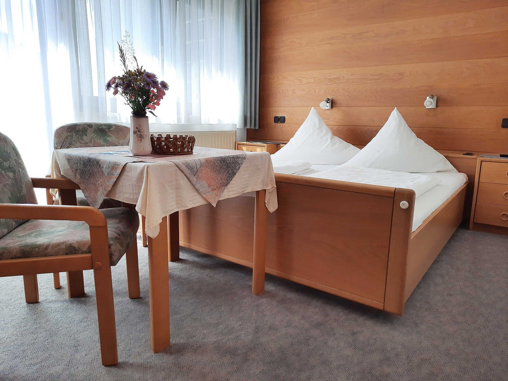 Ein Hotelzimmer mit einem Holzbett, einem kleinen Tisch mit Blumenvase, einem Sessel und einem Nachttisch.