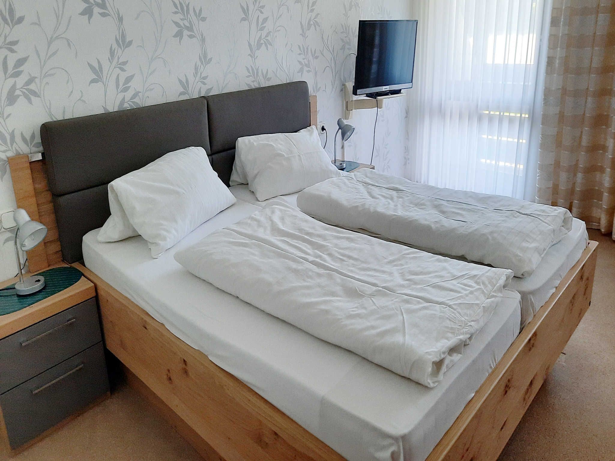 Ein Doppelbett mit weißer Bettwäsche, ein Nachttisch mit Lampe und ein an der Wand montierter Fernseher.