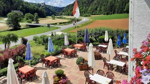 A terrace with outdoor seating, a flag, and a view of a road and forest. - Café-Pension Krems - Double room (Waischenfeld-Heroldsberg-Tal)