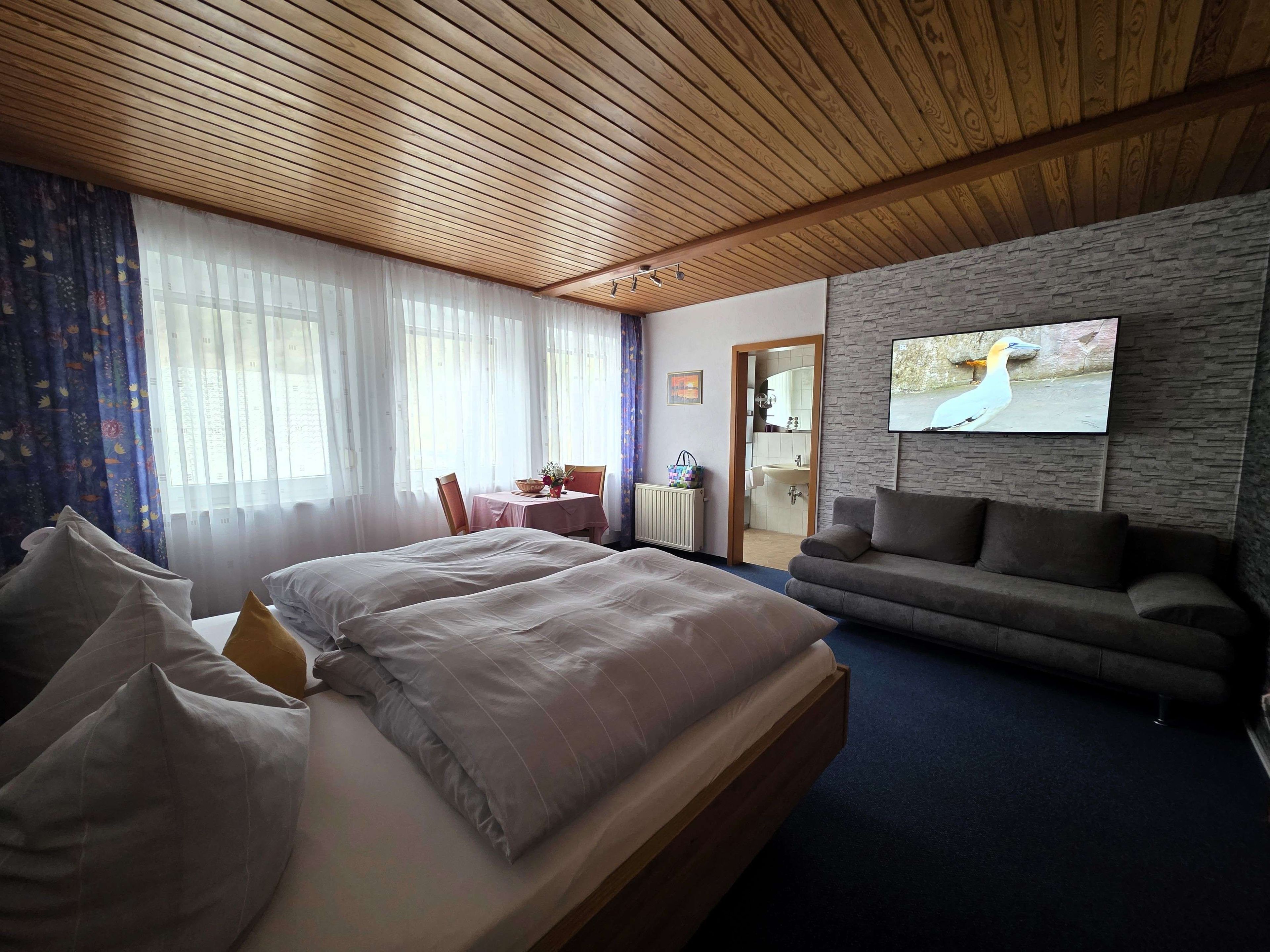 Ein Hotelzimmer mit Holzbalkendecke, einem Bett mit Kissen, einer Couch, einem an der Wand befestigten Fernseher und einem durch eine offene Tür einsehbaren Badezimmer.
