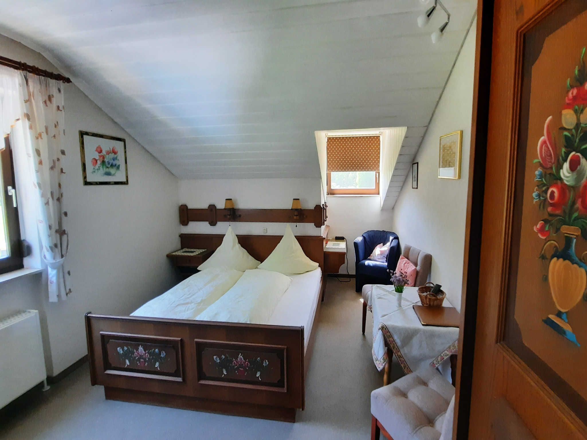 Une chambre à coucher avec un lit en bois, une petite table sur laquelle est posé un vase, une chaise et une fenêtre avec des rideaux.