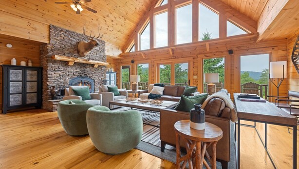 A spacious living room with a stone fireplace, wooden ceiling, and large windows offering a view of the outdoors. - Spacious Cabin in the Mountains (Banner Elk)