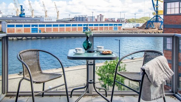 Parvekkeella on kaksi rottinkituolia, pieni pyöreä pöytä ja maljakko. Näköalasta avautuu vesistö ja taustalla näkyy teollisuusrakennuksia.
