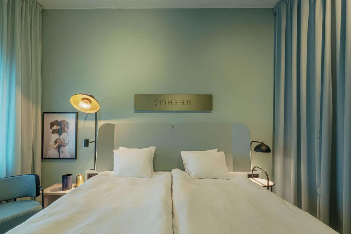 Ein Doppelbett mit weißer Bettwäsche, ein Nachttisch mit Lampe, ein gerahmtes Bild an der Wand und ein Schild mit der Aufschrift "(T) HIER".