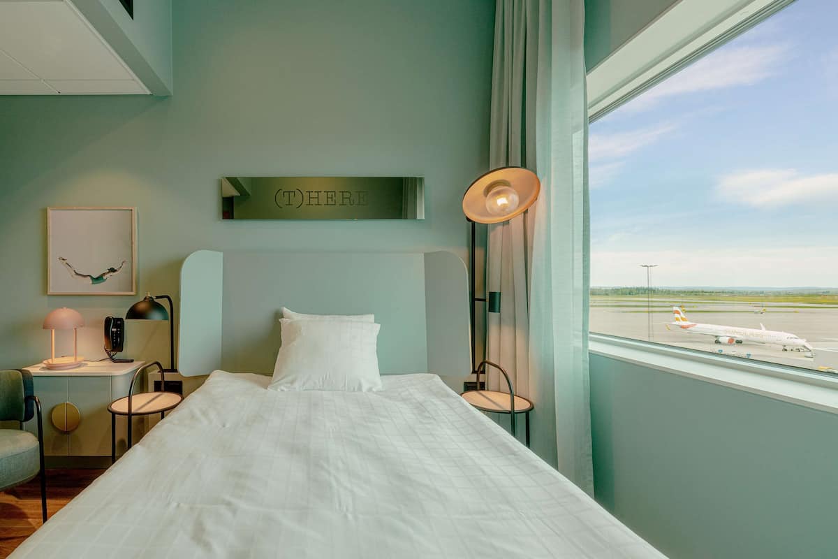Ein Hotelzimmer mit einem großen Bett, einem Schreibtisch mit Stuhl, einer Lampe, einem gerahmten Bild an der Wand und einem Fenster mit Blick auf ein Flugzeug am Rollfeld.