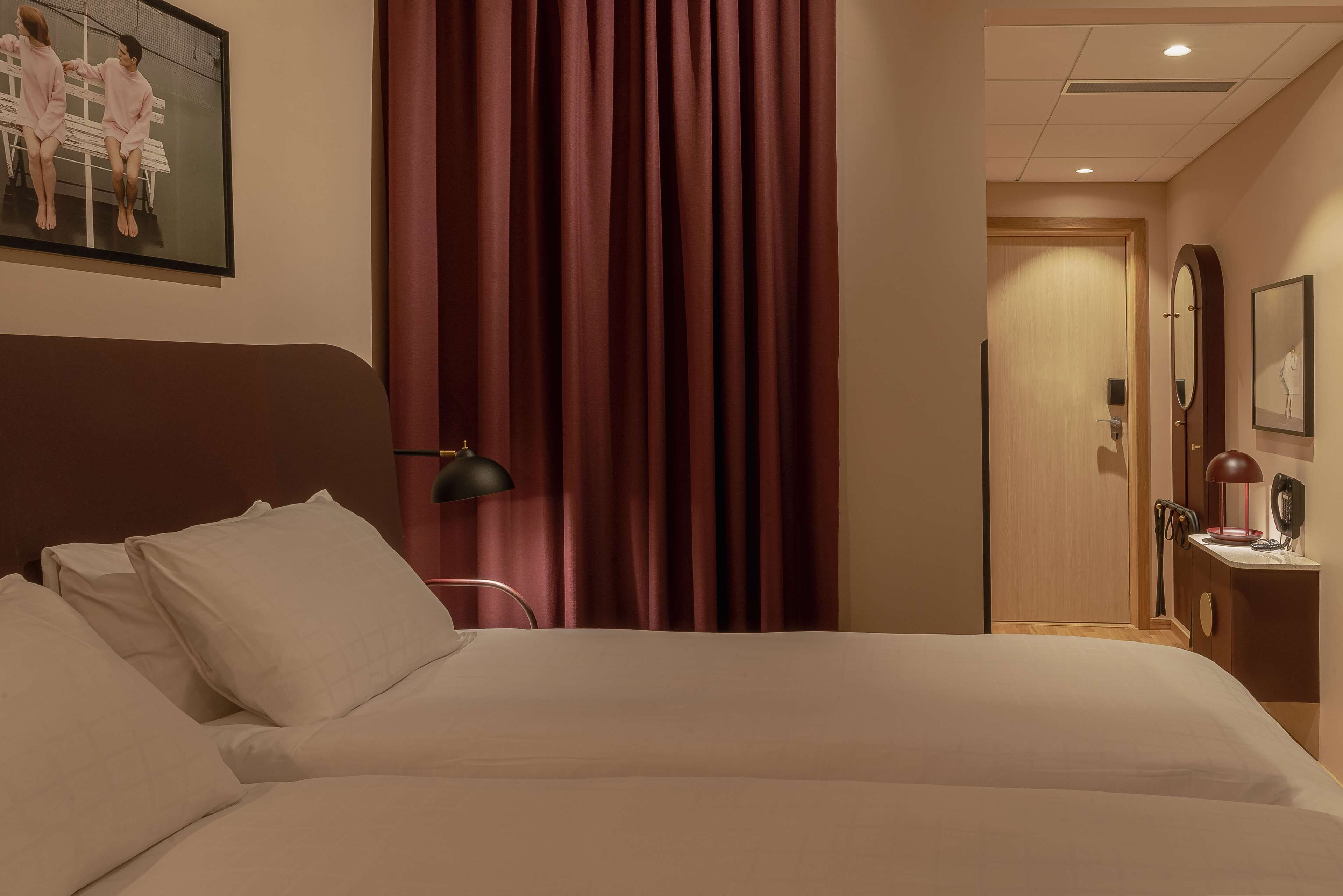 Ett hotellrum med en stor säng, ett skrivbord med en lampa, en spegel och en dörr som leder till ett annat rum.