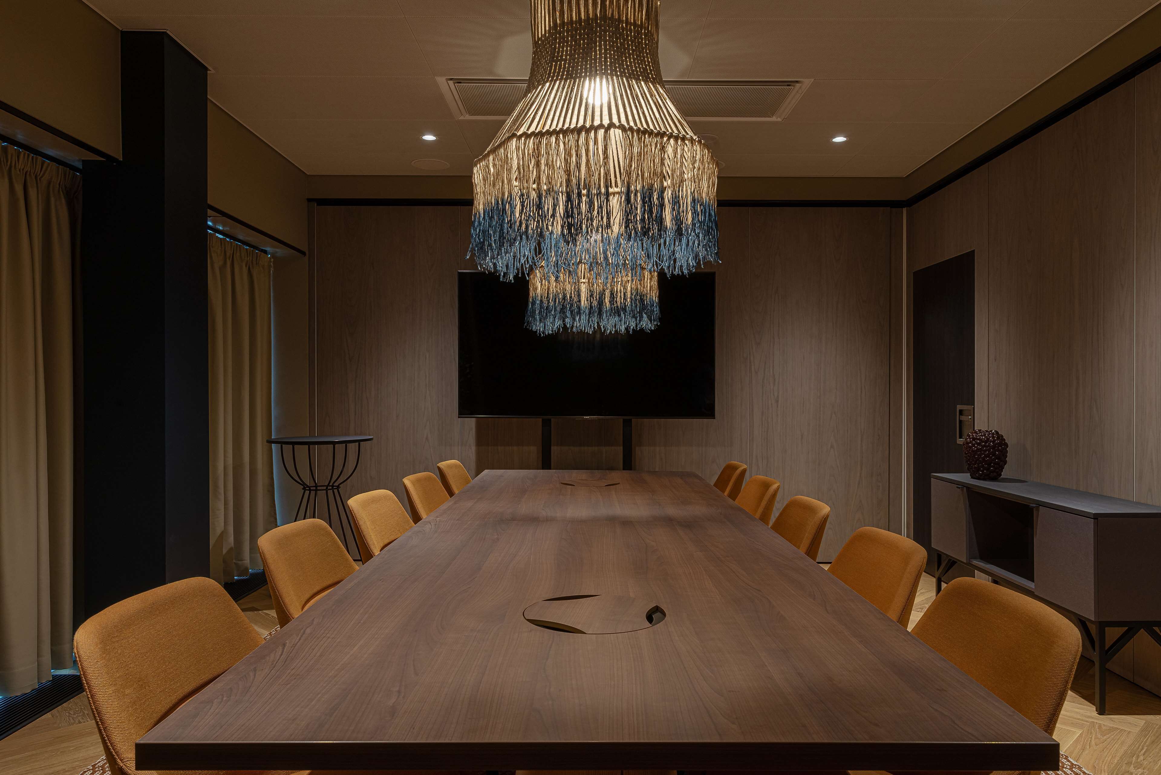 Un salón de conferencias con una larga mesa de madera, sillas anaranjadas, una lámpara de araña grande, un televisor de pantalla plana y un mueble pequeño.