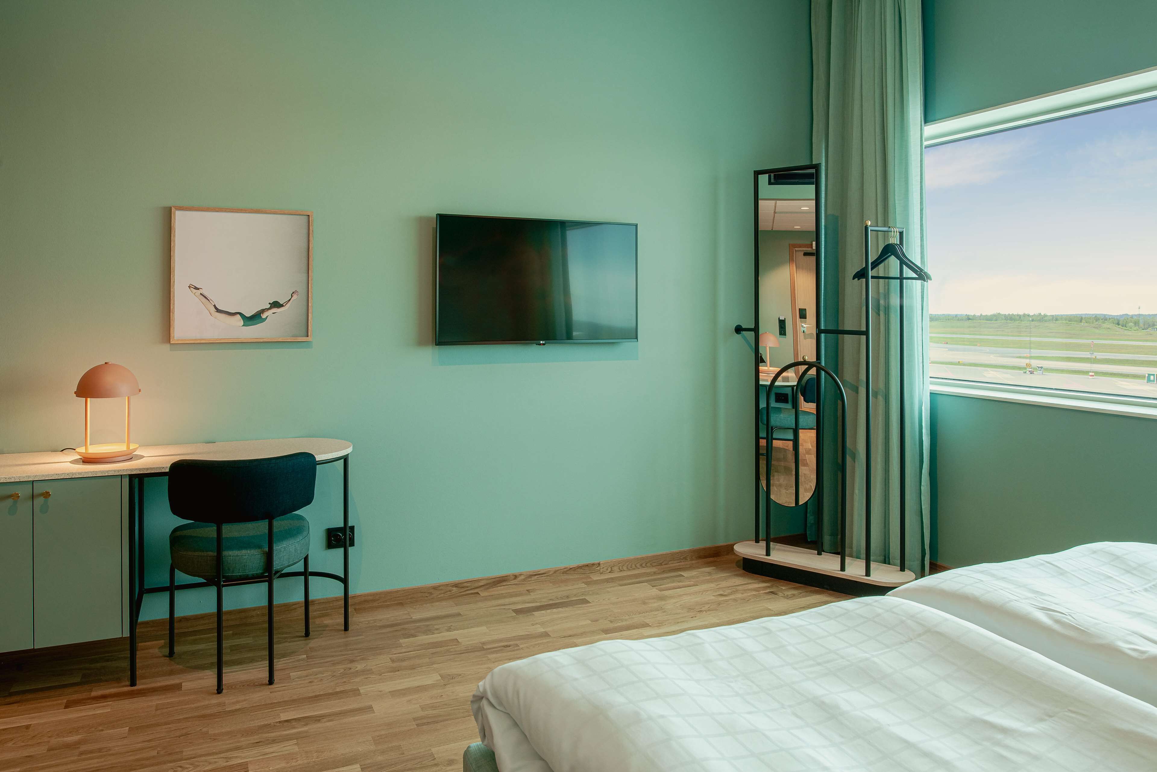 A hotel room with a bed, a desk with a chair, a TV, a framed picture, and a window with a view of the airport.