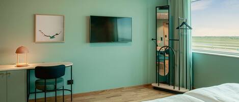 A hotel room with a bed, a desk with a chair, a TV, a framed picture, and a window with a view of the airport.