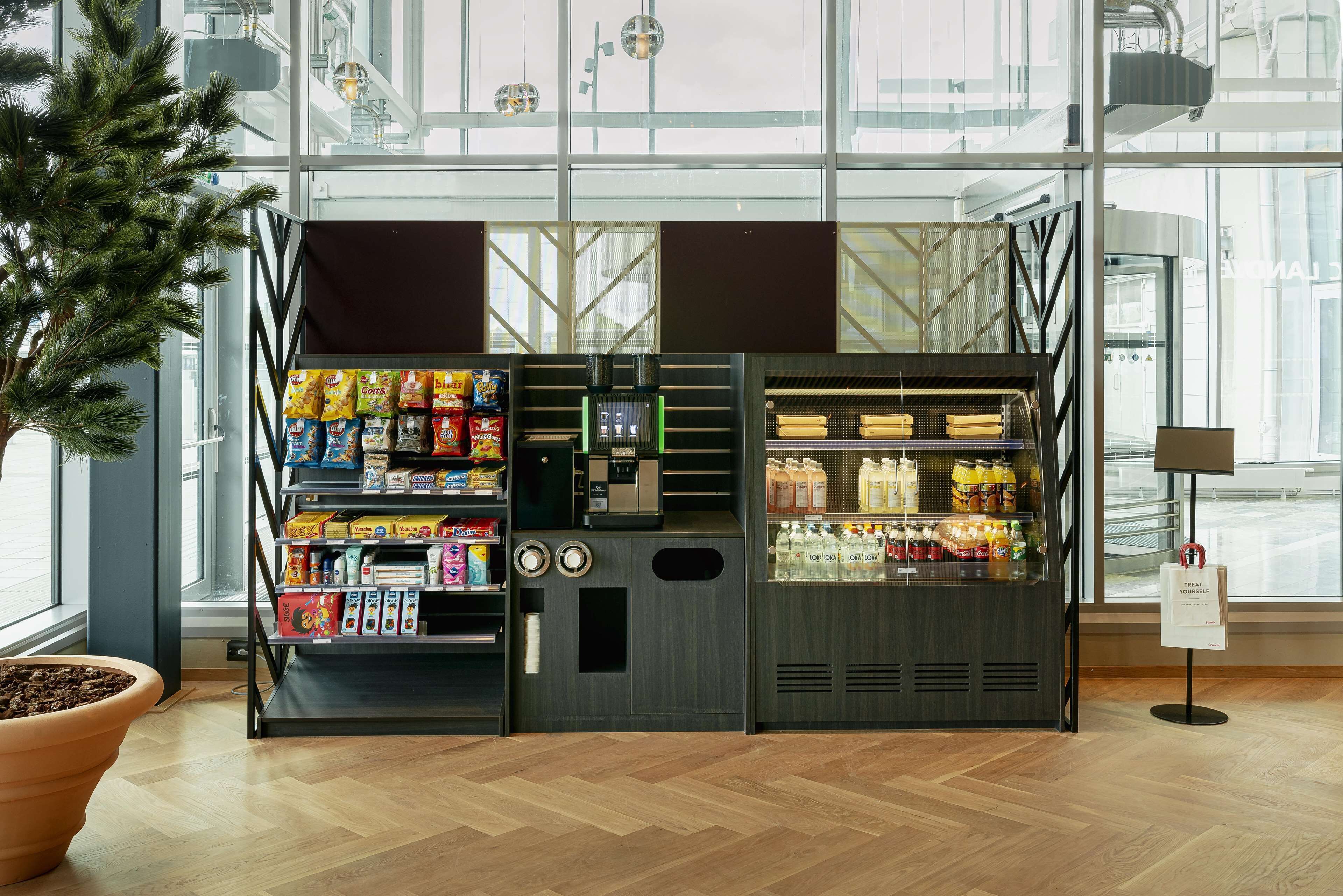 En automattankstation med snacks och drycker, placerad i en modern inomhusmiljö med stora fönster och en krukväxt.