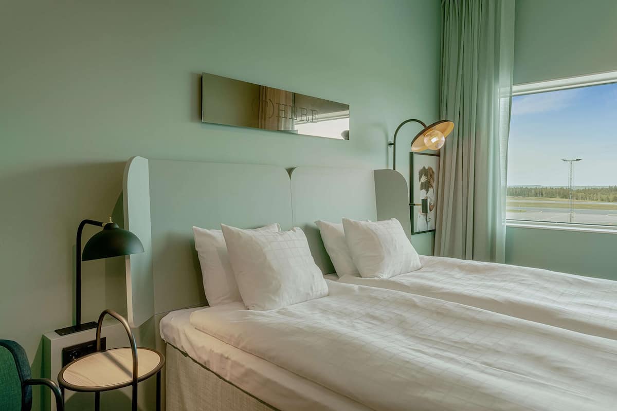 Ein Hotelzimmer mit einem großen Bett, einem Sessel, einer Lampe und einem Fenster mit Blick auf das Flughafengelände.