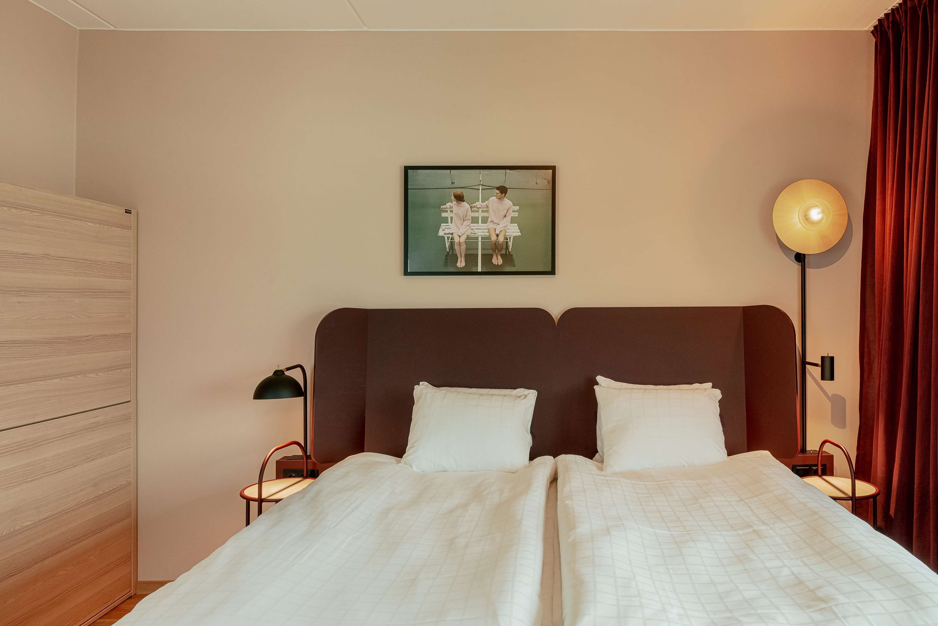 Uma cama de casal com roupa de cama branca, cabeceira, duas mesas de cabeceira com abajures, um quadro na parede e um guarda-roupa de madeira.