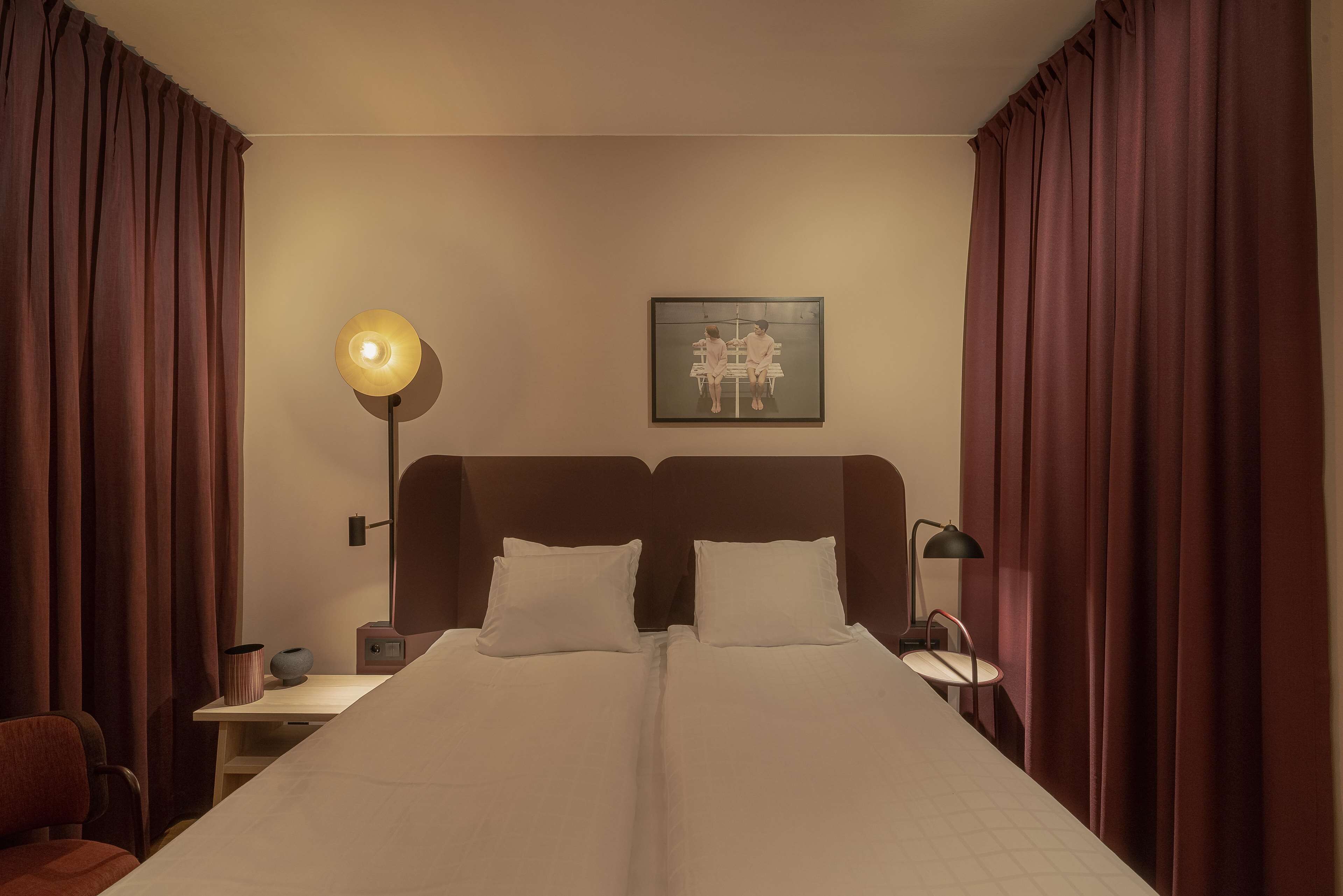 Una cama doble con ropa de cama blanca, una mesita de noche con lámpara, una obra de arte enmarcada en la pared y cortinas marrones.