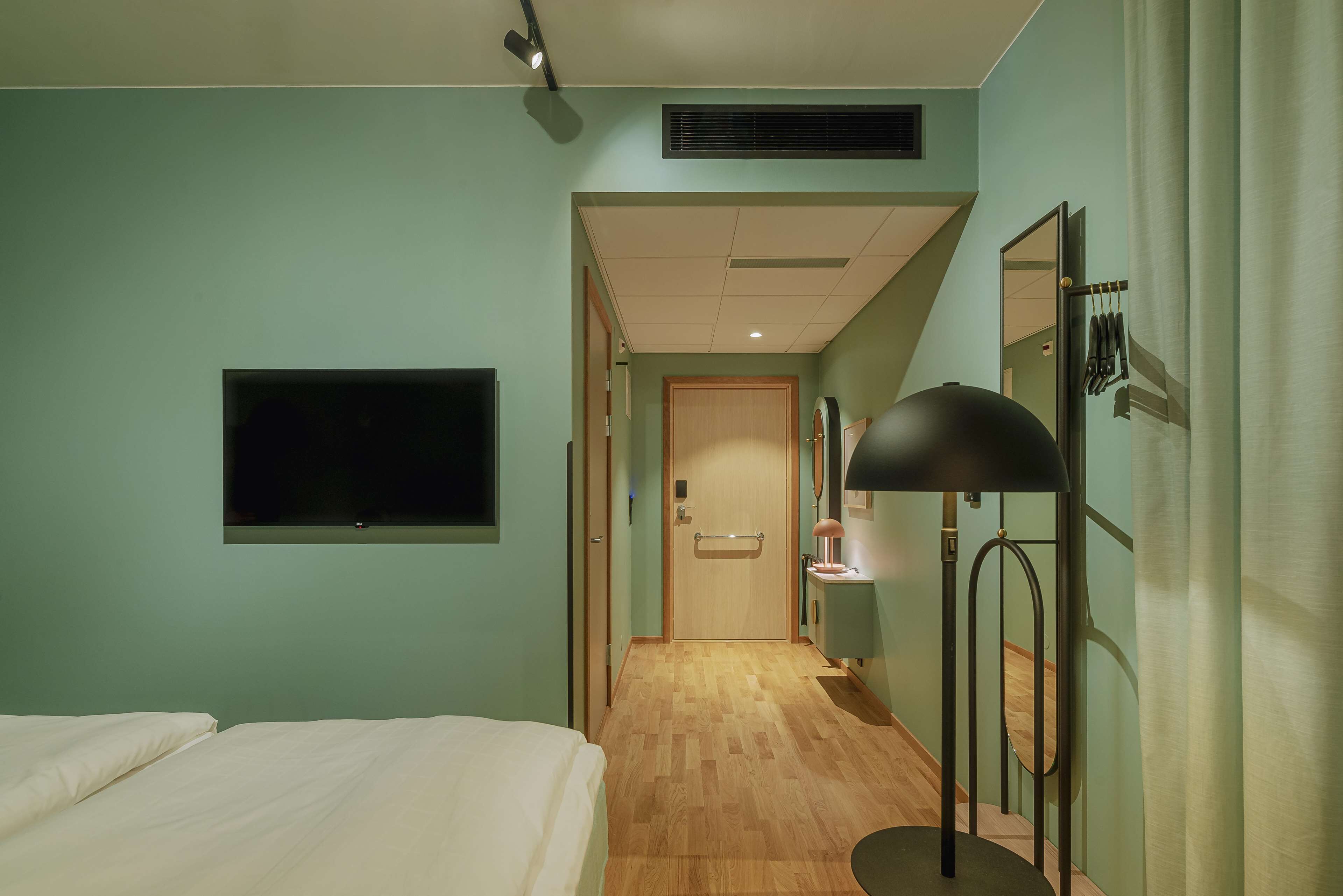 A hotel room with a bed, a TV mounted on the wall, a desk with a lamp, and a mirror.
