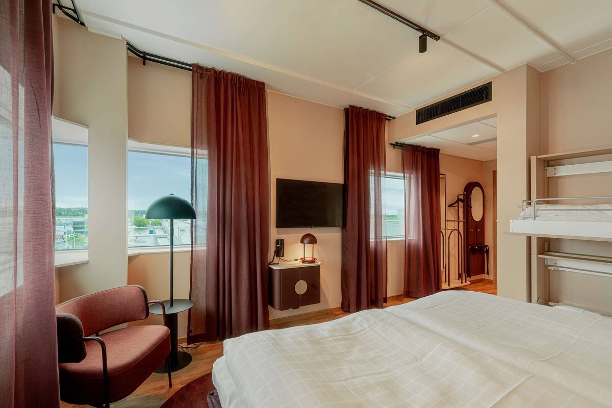 Ein Hotelzimmer mit einem Bett, einem Schreibtisch mit Lampe, einem Sessel, einem Fernseher und einer Gitarre.