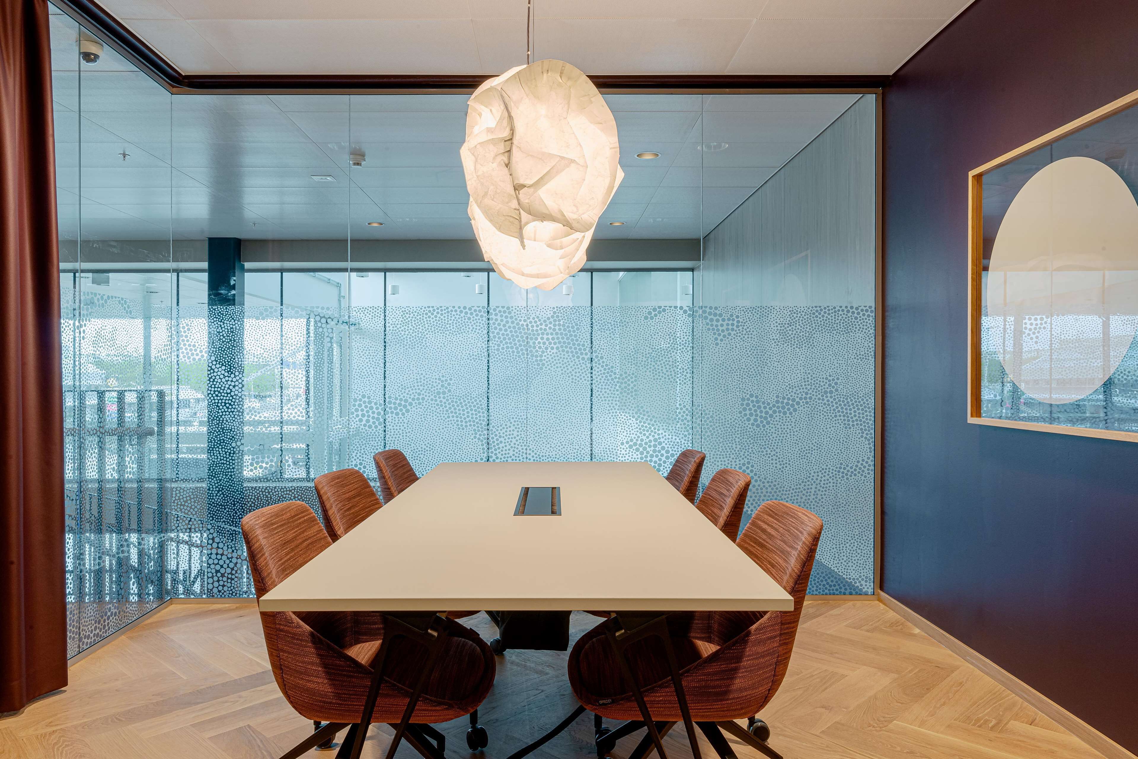 A conference room with a large table, eight chairs, a hanging light fixture, and a patterned glass wall.