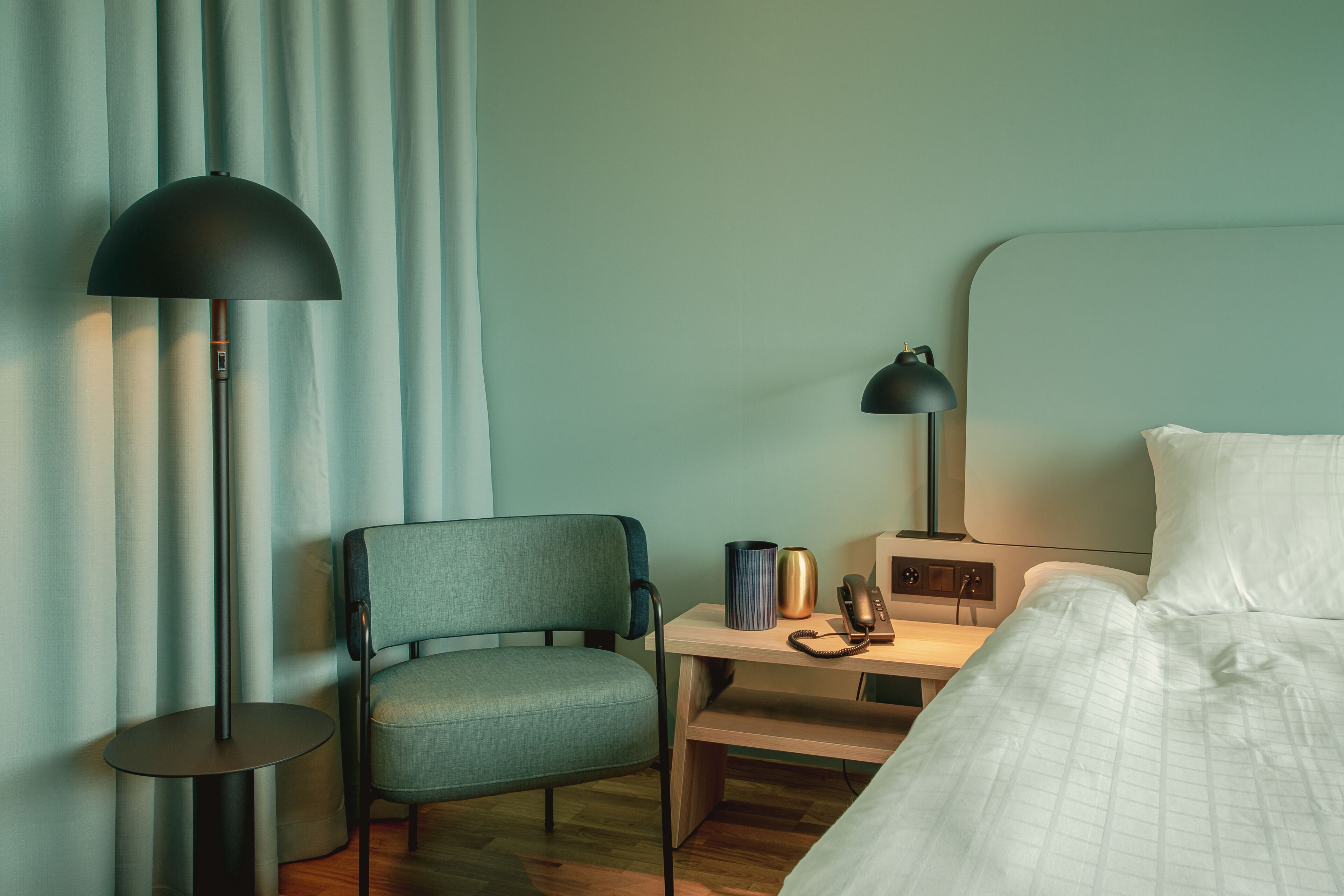 A hotel room with a bed, a desk with a lamp, a chair, and a bedside table with a phone and a vase.