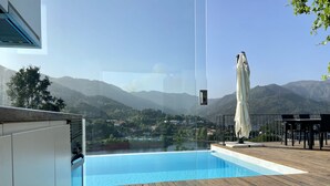 Un área de piscina moderna al aire libre con un deck de madera, piscina cubierta con vidrio y vistas a un paisaje montañoso.