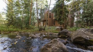 A rustic cabin surrounded by a forest with a stream in front.