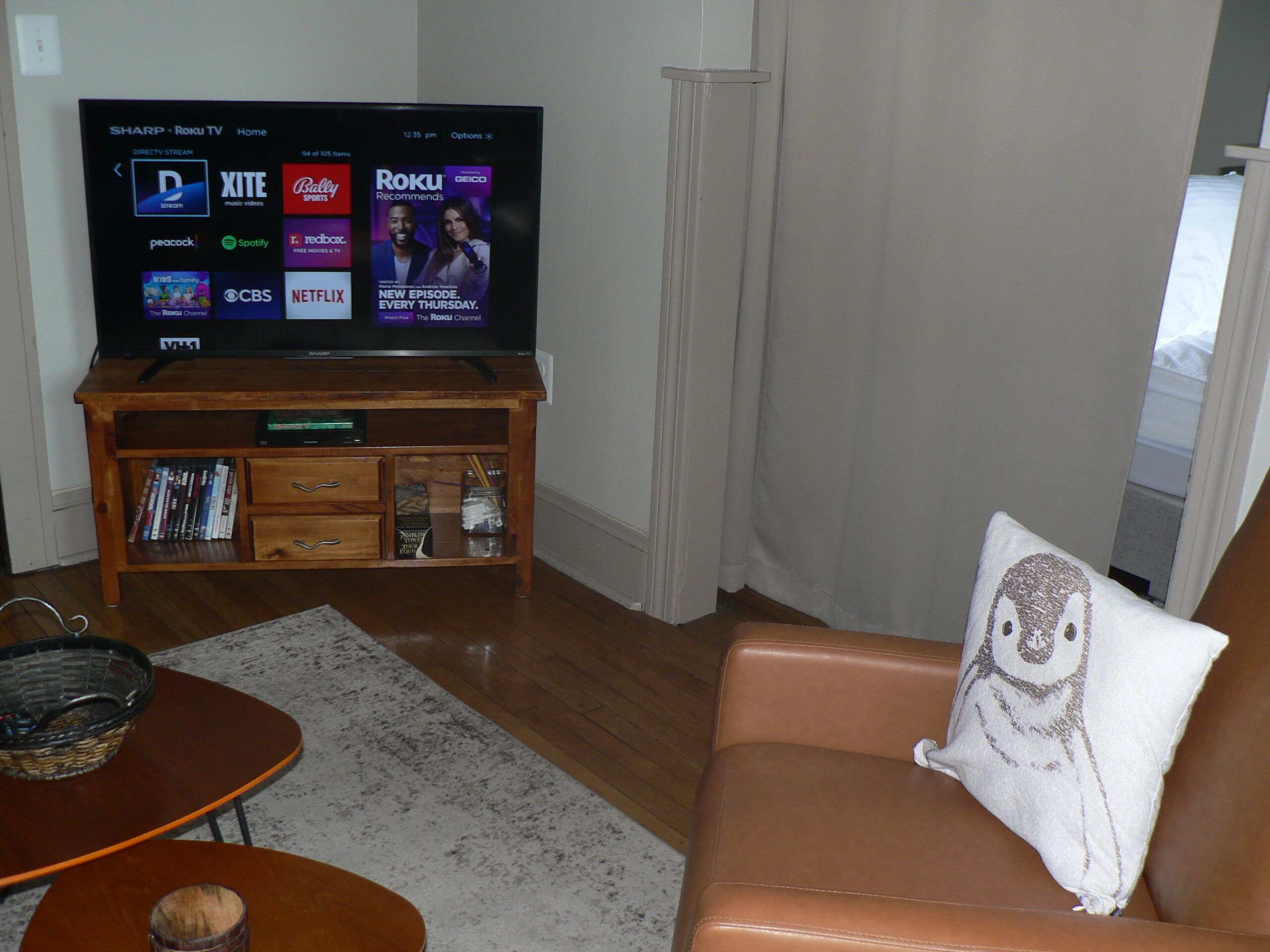 Ein Wohnzimmer mit einem Flachbildfernseher, der Streaming-Dienste anzeigt, einer Holzunterhaltungsecke mit Büchern und einem braunen Ledersofa mit Kissen.