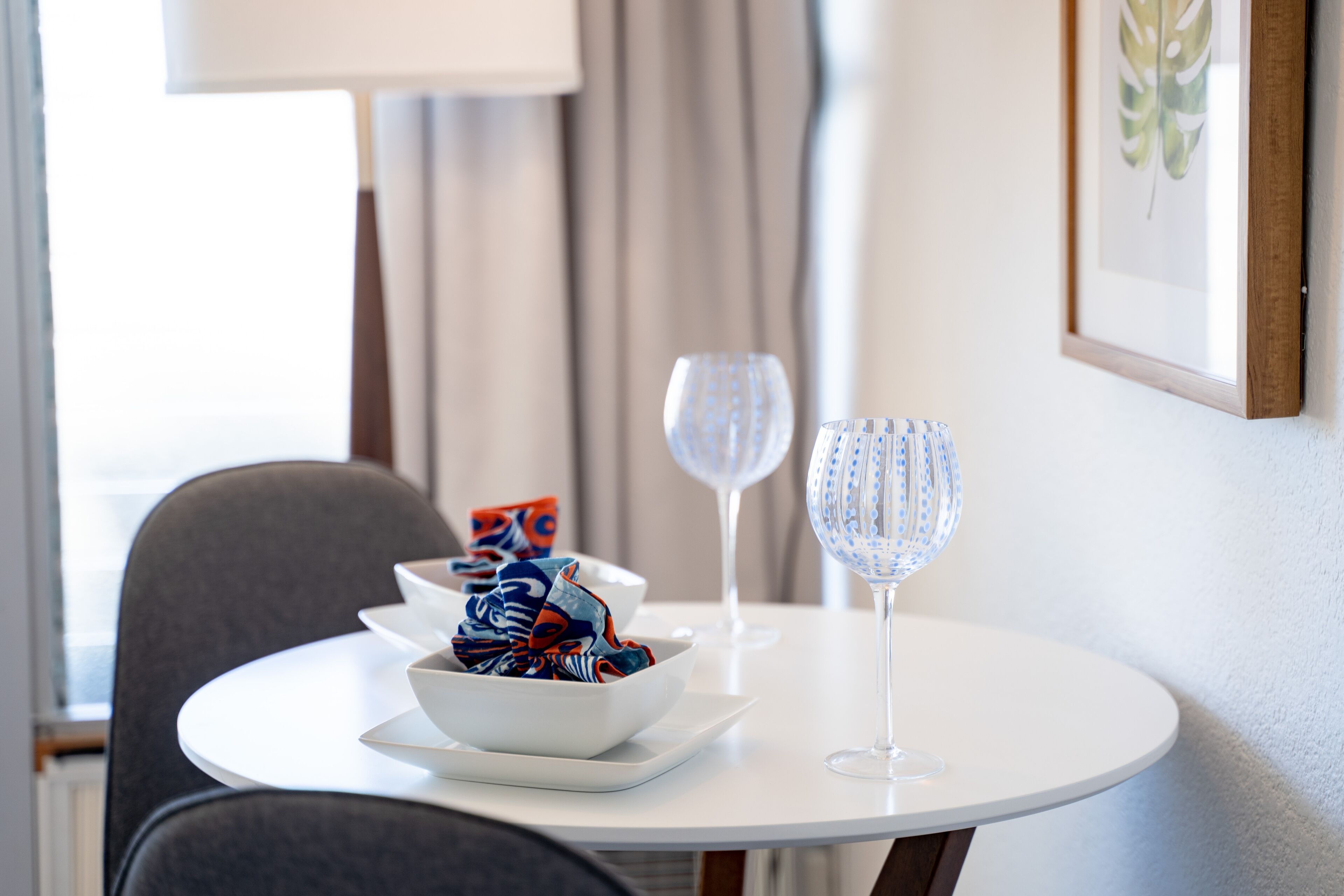 Une table de salle à manger ronde, recouverte d’une nappe blanche, avec un saladier de fruits et deux verres à vin.