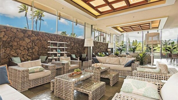 A spacious living area with wicker furniture, a stone wall, and large windows offering a view of palm trees.