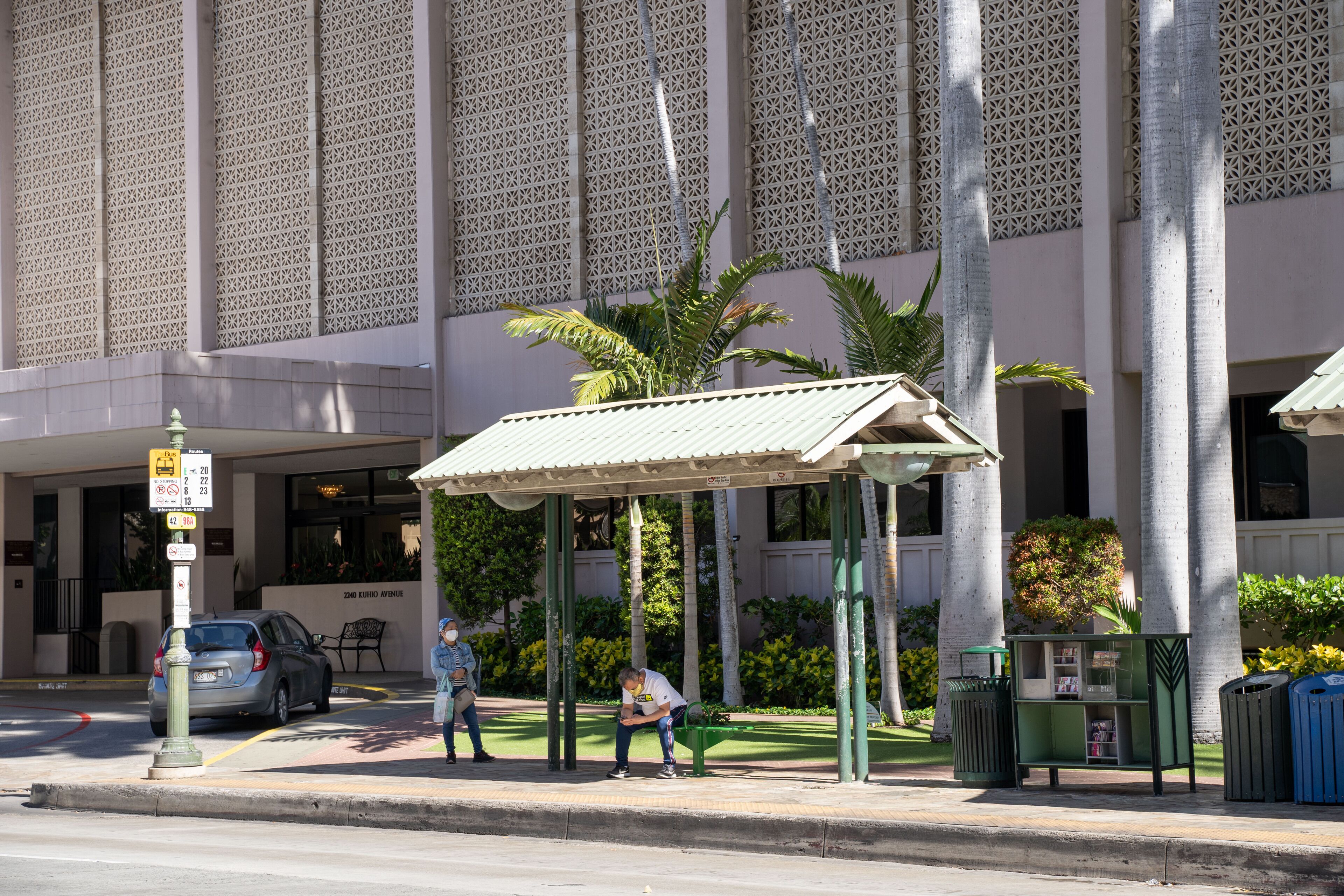 A building with a patterned facade, a covered waiting area with palm trees, and a street with cars and pedestrians.
