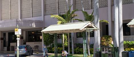 A building with a patterned facade, a covered waiting area with palm trees, and a street with cars and pedestrians.