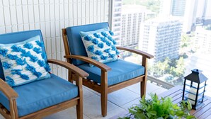 Two wooden chairs with blue cushions and blue and white patterned pillows, a potted plant, and a black lantern on a balcony with a city view.