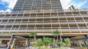 A multi-story hotel with a central entrance, balconies, and a landscaped area with plants.