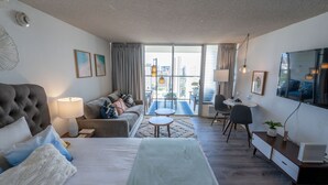 A hotel room with a bed, a sofa, a desk, a chair, a small table, and a view of the city through large windows.