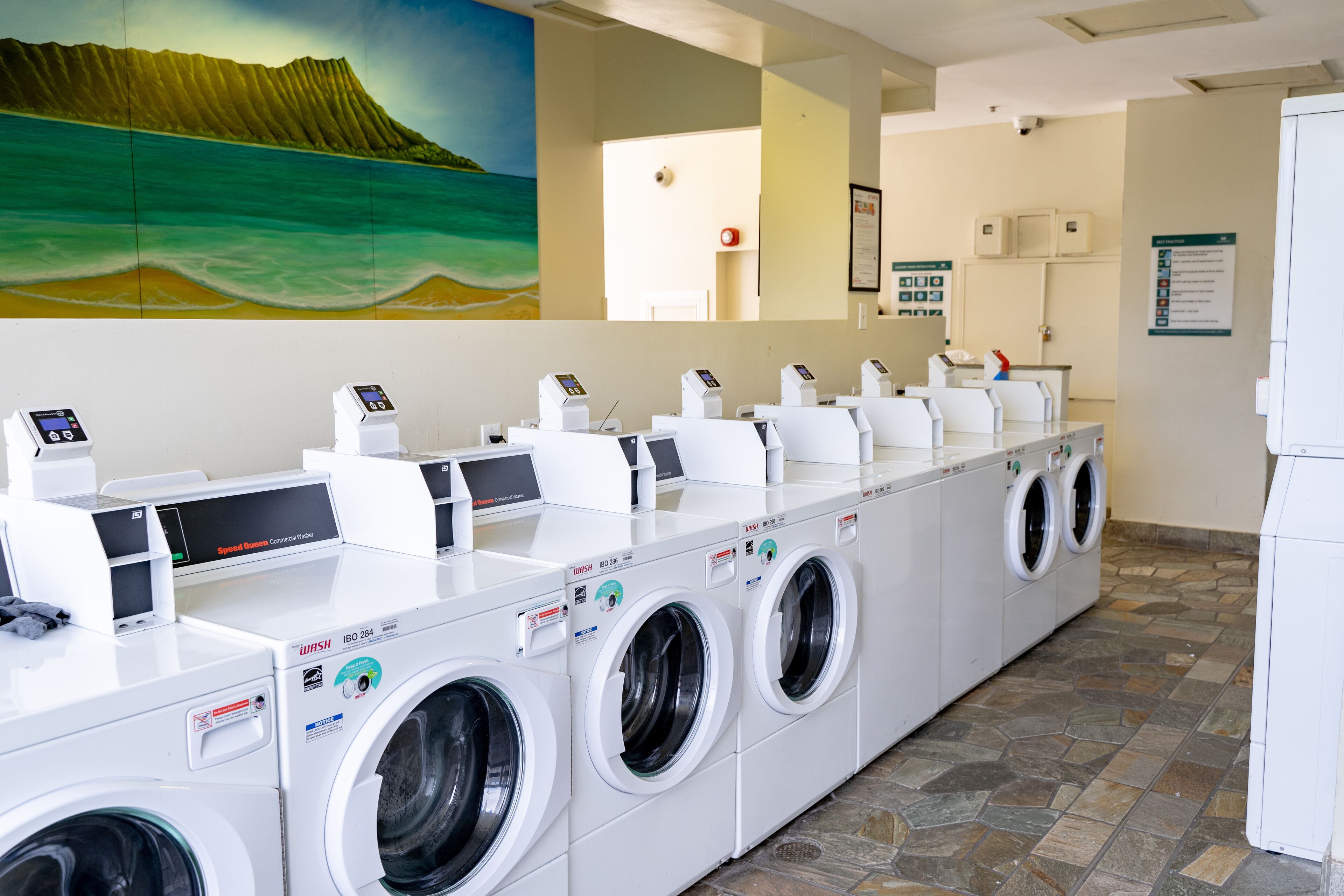 Een wasserette met meerdere wasmachines en drogers, een muurschildering van een strandlandschap en een betegelde vloer.