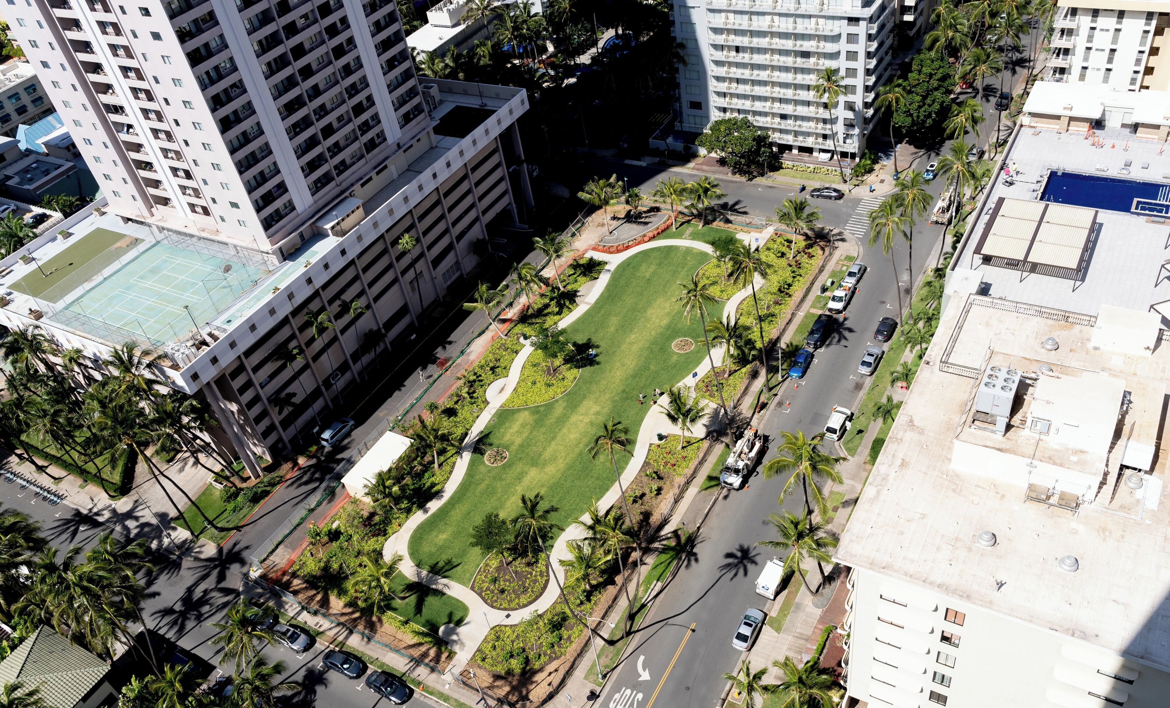 An aerial view of a modern urban area with high-rise buildings, a central landscaped park, and a road with vehicles.