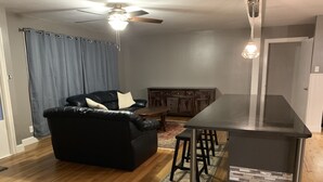 Ein Wohnzimmer mit einem schwarzen Ledersofa, einem Holzesstisch und einer Küchenzeile mit schwarzer Arbeitsplatte und Hockern.